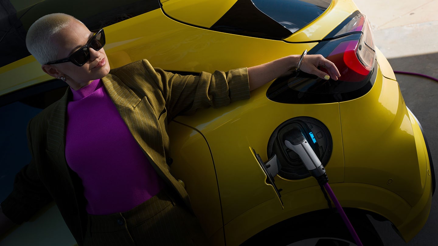 Een vrouw leunt tegen een gele elektrische Ford Capri die is aangesloten op een oplaadstation voor elektrische voertuigen.